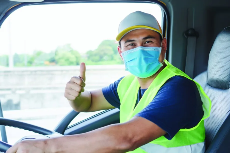Delivery driver wearing a face mask and high-visibility vest gives a thumbs-up from the cab of a truck.