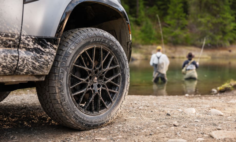 An SUV's tire is showcased beside a calm body of water, highlighting the vehicle's rugged design in a natural setting.