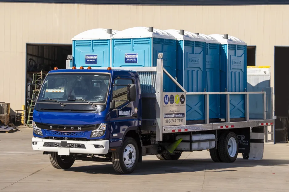 Battery electric truck with blue cab and flatbed hauling portable toilets.