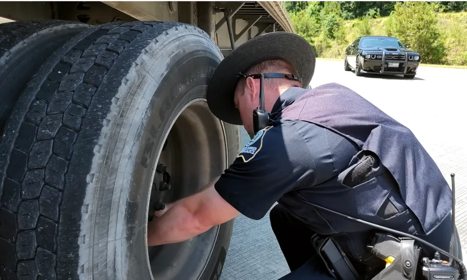 A state police officer inspects the wheels of a trailer with his patrol car parked in the background.