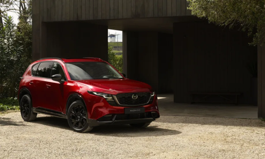 Front three-quarter view of a red 2026 Mazda CX-5 parked on a gravel driveway in front of a modern, dark wood building, featuring black wheels and Mazda's Signature Wing grille.