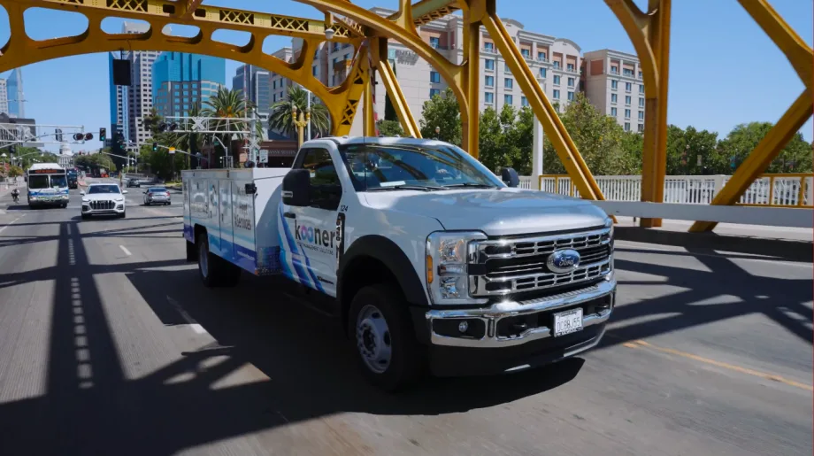 A white service truck driving across a yellow bridge with city skyline in the background.
