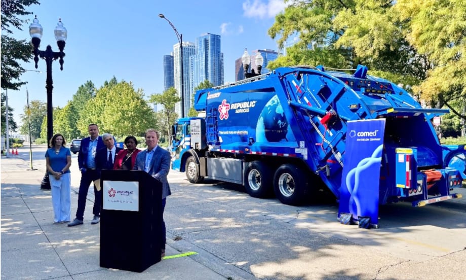 Group of officials during an event showcasing a blue Mack LR Electric refuse truck, one speaker is at the podium talking and others are in the background.