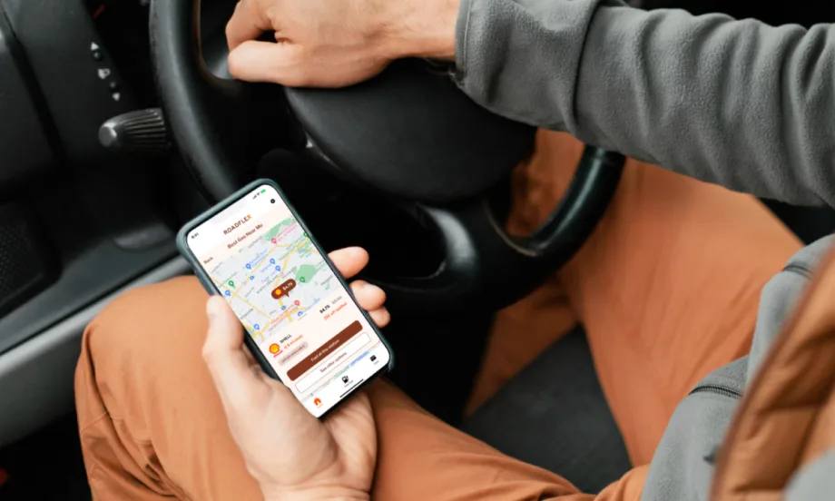 Driver holding a phone with the RoadFlex fuel management app open, showing a map with gas stations and real-time prices inside a vehicle.