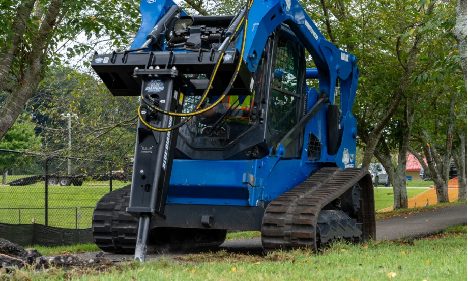 Blue skid steer using a hydraulic breaker to demolish paved sidewalk in a park setting.