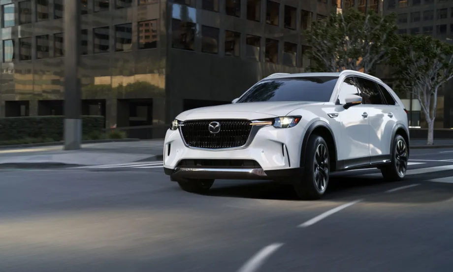 A white 2026 Mazda CX-90 crossover SUV driving through a city street, showcasing its front grille, LED headlights, and modern exterior design.