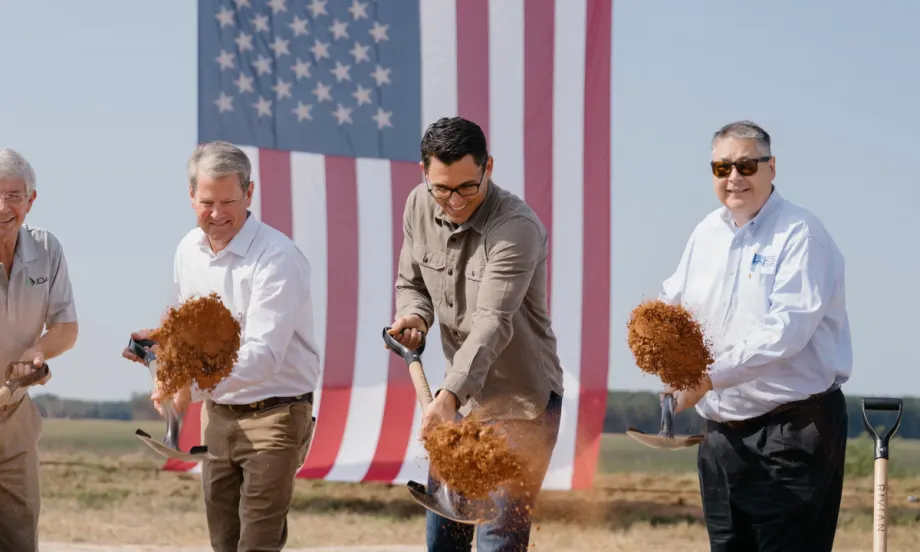 Rivian CEO RJ Scaringe, Georgia Governor Brian Kemp, and other officials shovel dirt during the groundbreaking ceremony, with a large American flag in the background.