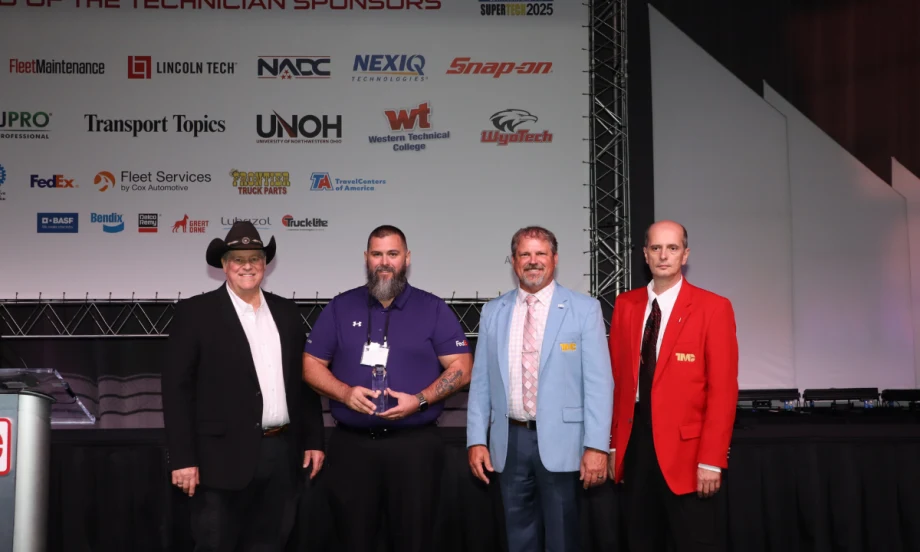 Matthew Sloan, winner of TMCSuperTech 2025, stands with event officials and holds his award during the recognition ceremony, with sponsor logos displayed on the backdrop.