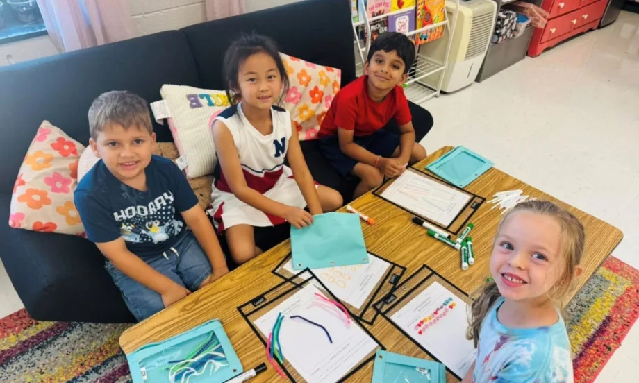 Four young students sit at a classroom table with worksheets, markers, and colorful pipe cleaners, smiling while engaging in a STEM activity.