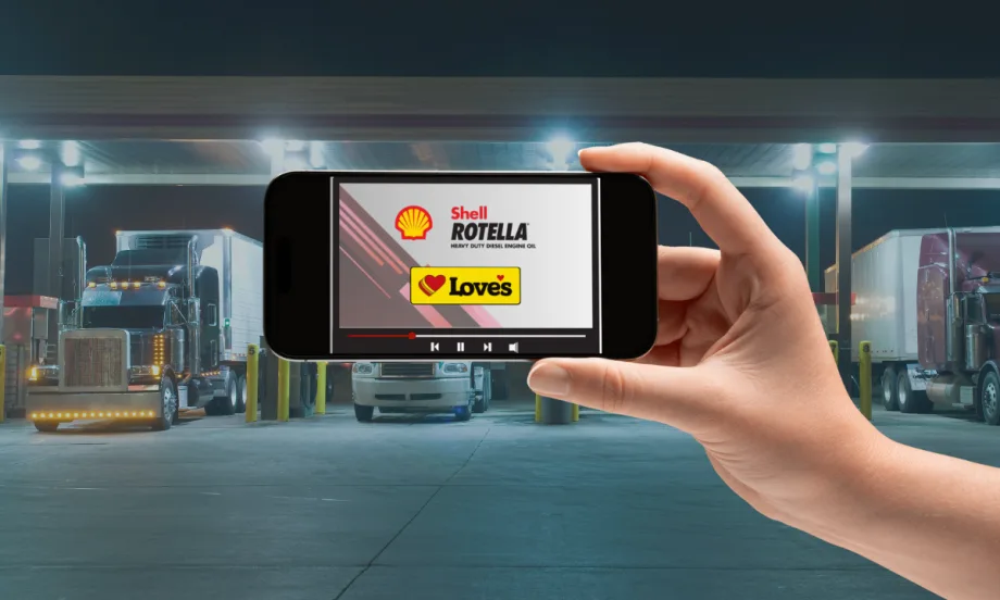 Hand holding a smartphone displaying a Shell Rotella and Love’s Travel Stops video screen, with semi-trucks parked at a brightly lit fuel station in the background.