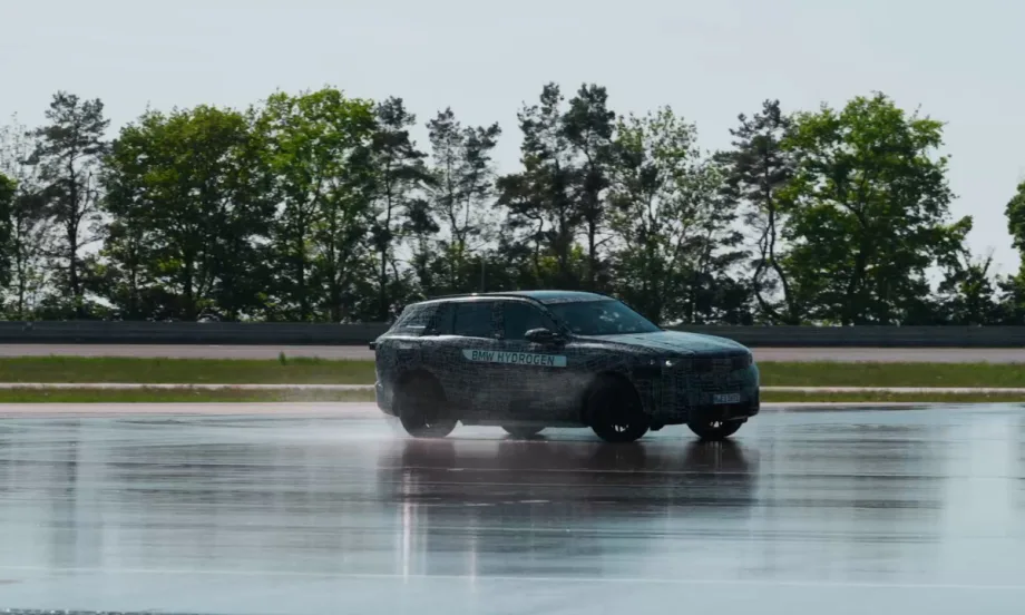 Camouflaged BMW iX5 Hydrogen SUV driving on a wet test track, kicking up water spray, with trees and a racetrack barrier in the background.