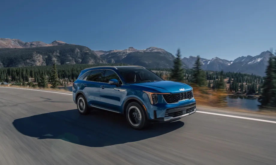 Blue 2026 Kia Sorento Hybrid driving on a mountain highway with trees and water in the background.
