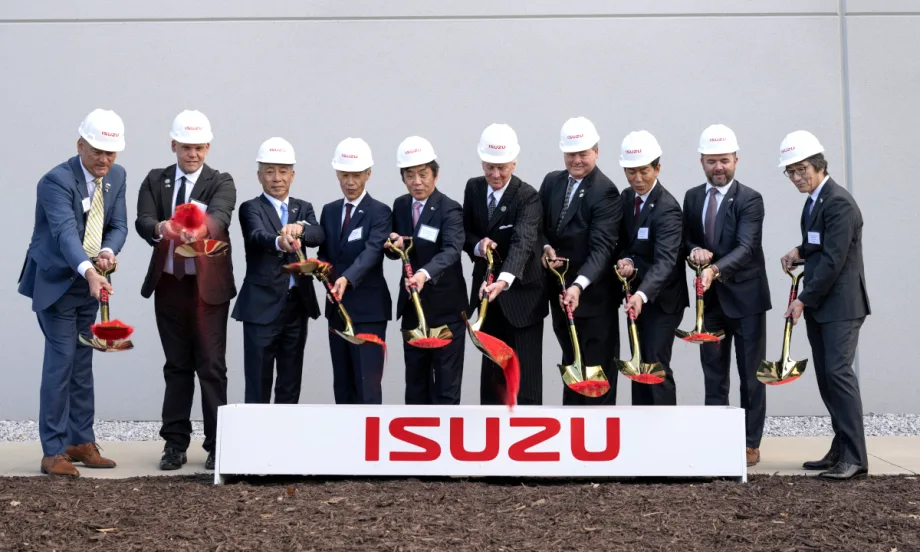 Business men stand behind a box with Isuzu logo and shovel dirt onto the ground.