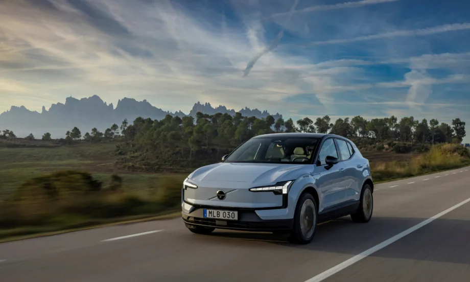 A light blue 2026 Volvo EX30 electric SUV driving on a rural highway with mountains and trees in the background under a clear sky.