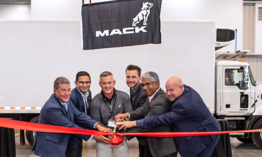 Group of businessmen cut ribbon in front of a white box truck with a Mack Trucks sign on the side.