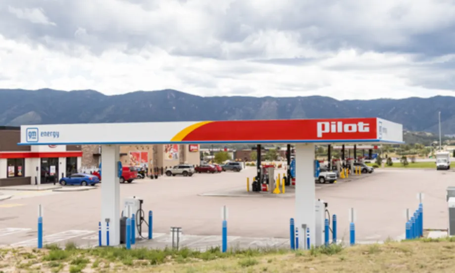 A Pilot Travel Center with GM Energy and EVgo fast-charging stations installed under the canopy, with fuel pumps and vehicles in the background against a mountain landscape.