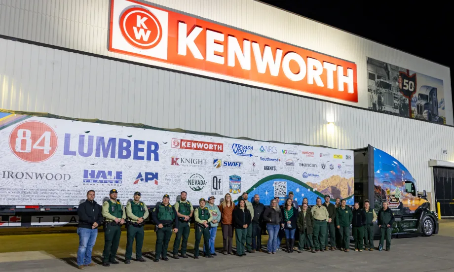 Group of people stand in front of a tractor trailer parked outside a building with large Kenworth sign