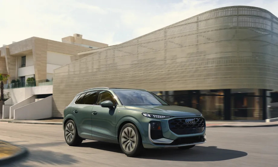 Green 2026 Audi Q3 compact SUV driving on a city street past a modern building in daylight.
