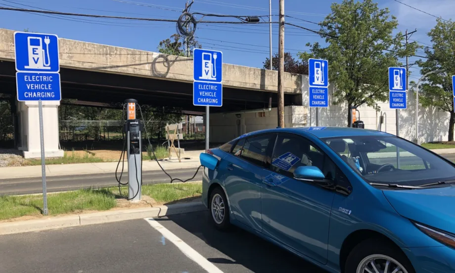 Blue electric car plugged into an EV charging station beneath an overpass, with multiple “Electric Vehicle Charging” parking signs.