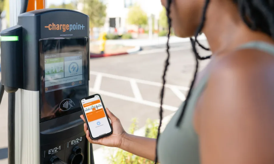 A driver uses the ChargePoint app on a smartphone next to a ChargePoint EV charging station.