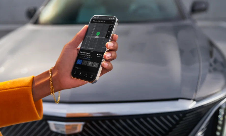 A driver holds a smartphone displaying a GM vehicle app with an Electrify America charging station map, with an electric vehicle parked in the background.