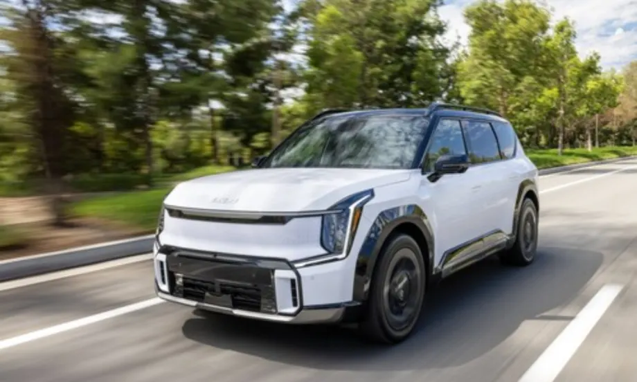 A white 2026 Kia EV9 electric SUV driving on a tree-lined roadway.