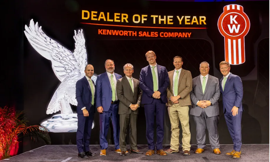 Group of men in business attire on a stage during an award ceremony with background that says Dealer of the Year and a Kenworth logo.