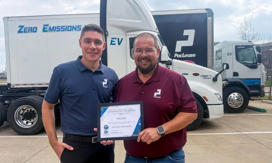 Two men stand together holding a certificate award with large commercial trucks in the background.