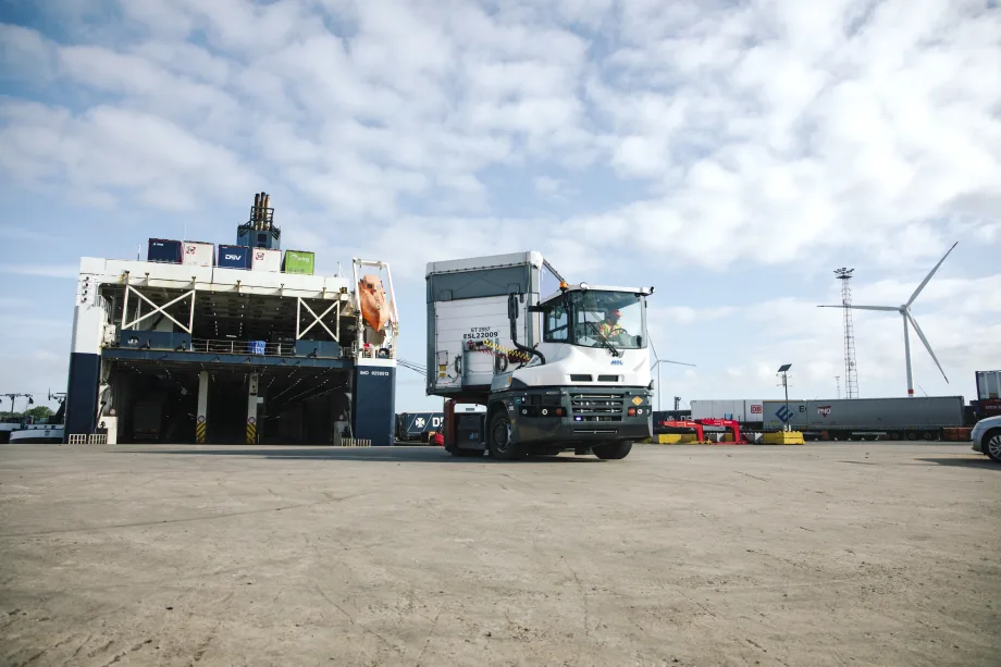 Terminal truck hauling a container trailer with a cargo ship and windmill in the background.