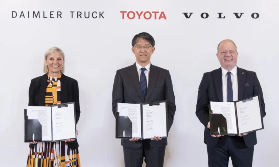 Woman and two men standing holding paperwork agreements they have signed with logos for Daimler Truck, Toyota, and Volvo over their heads.