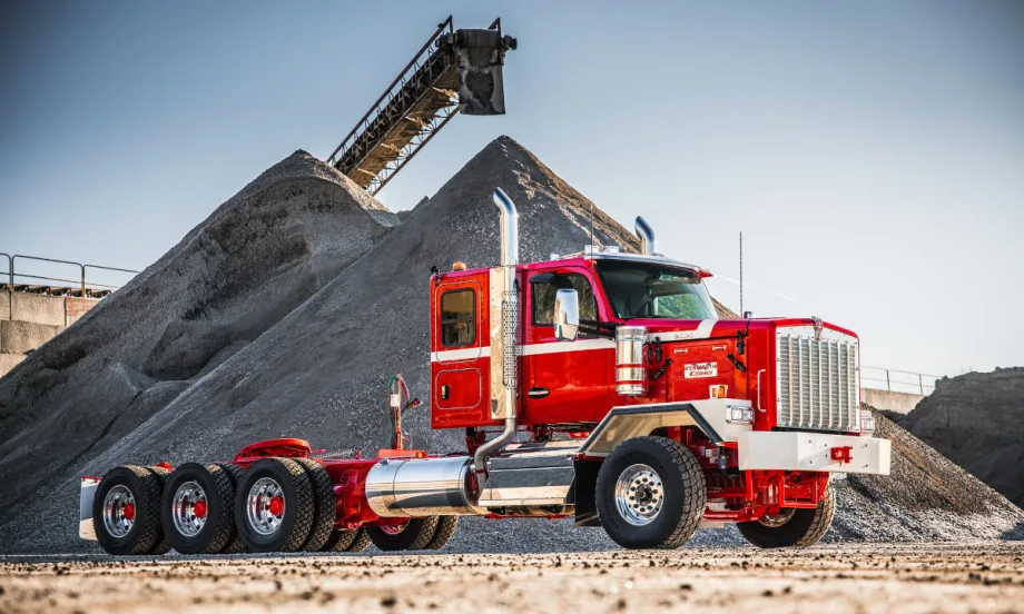 Red Kenworth truck parked sideways with large mound of gravel in background.