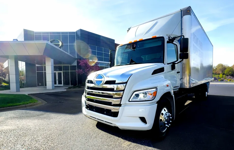 White box delivery truck parked outside a modern office building on a sunny day. The truck is photographed from the front angle, with chrome detailing reflecting sunlight and the building entrance visible in the background.