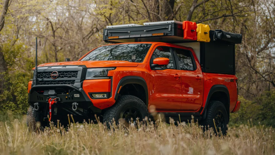 Orange Nissan Frontier PRO-4X overland build parked in a grassy wooded area with off-road tires, heavy-duty front bumper, winch, rooftop cargo storage, and camping accessories mounted above the truck bed.