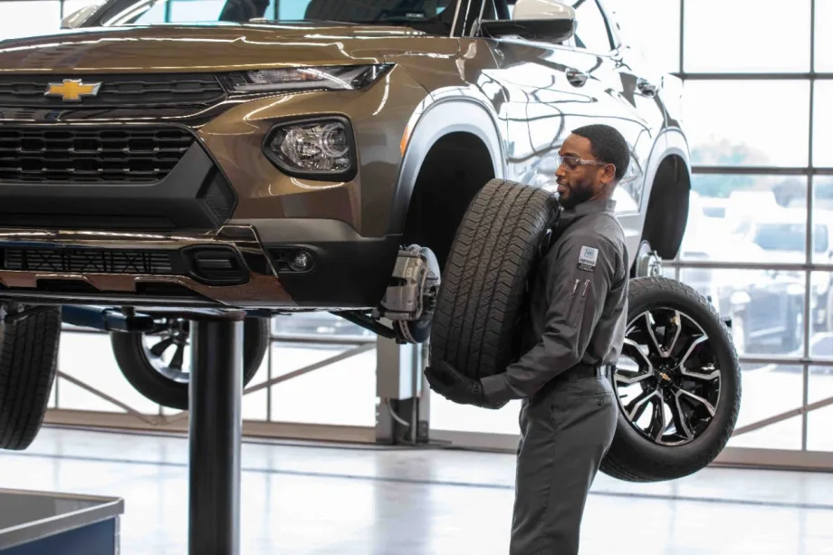 Automotive technician carrying a tire beside a Chevrolet SUV raised on a service lift inside a repair facility. Additional wheels and bright garage windows are visible in the background.