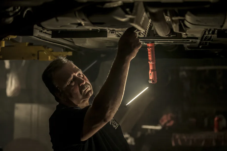 Mechanic working beneath a raised vehicle in a dimly lit garage, illuminated by a hanging Streamlight work light. The technician reaches toward the undercarriage while inspecting or repairing automotive components.