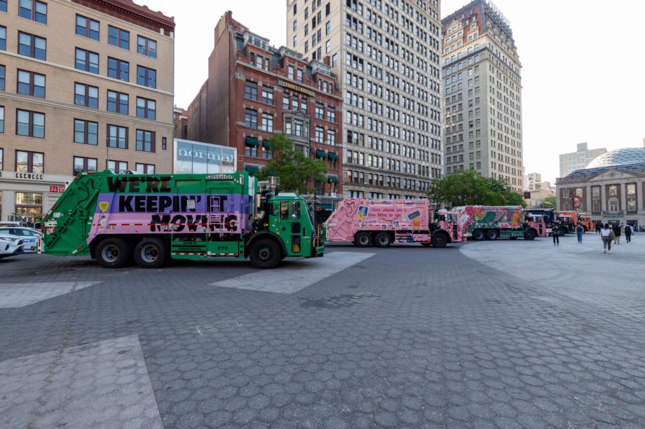 Line of brightly painted New York City sanitation trucks displayed in a public plaza, featuring colorful murals and messages. Tall city buildings and pedestrians surround the outdoor exhibition in an urban setting.