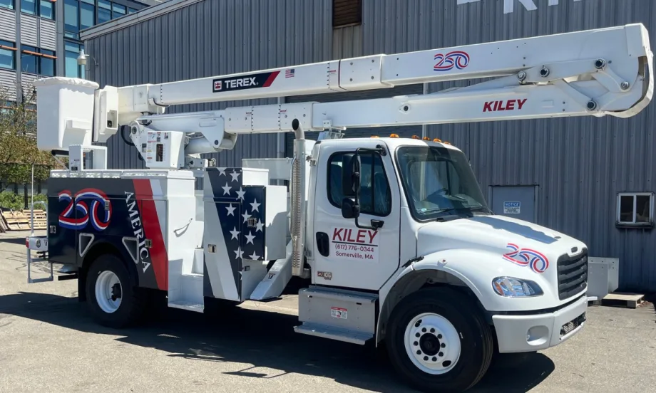 White Kiley utility truck equipped with a Terex aerial lift parked outside an industrial building. The truck features patriotic red, white, and blue graphics, including an American flag design and “250” anniversary emblems on the body and boom.