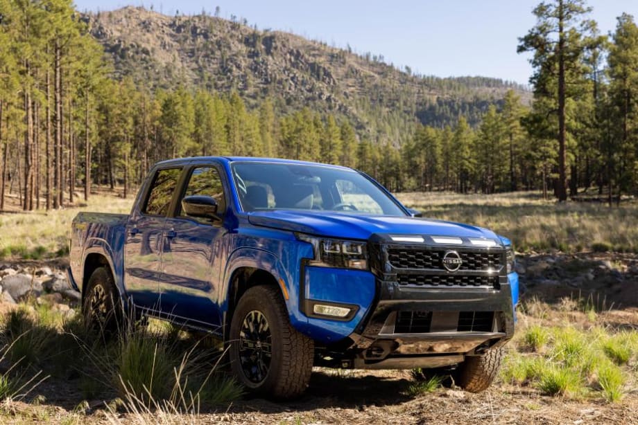 Blue Nissan Frontier pickup truck parked on rugged terrain in a forested mountain landscape. The four-door truck is shown from the front three-quarter angle, highlighting its off-road stance, black grille, and outdoor adventure setting.