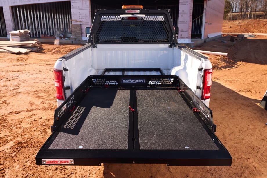Rear view of a white work truck at a construction site featuring a Weather Guard sliding truck-bed system extended outward, providing easy access to tools and equipment. A protective cab rack and unfinished building frame the background.