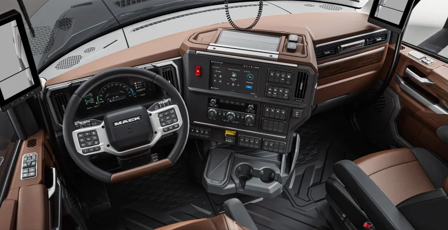 Driver’s-eye view of a Mack Granite truck interior featuring a digital instrument cluster, multifunction steering wheel, large touchscreen infotainment system, integrated controls, cup holders, and premium brown-and-black trim designed for comfort and productivity.