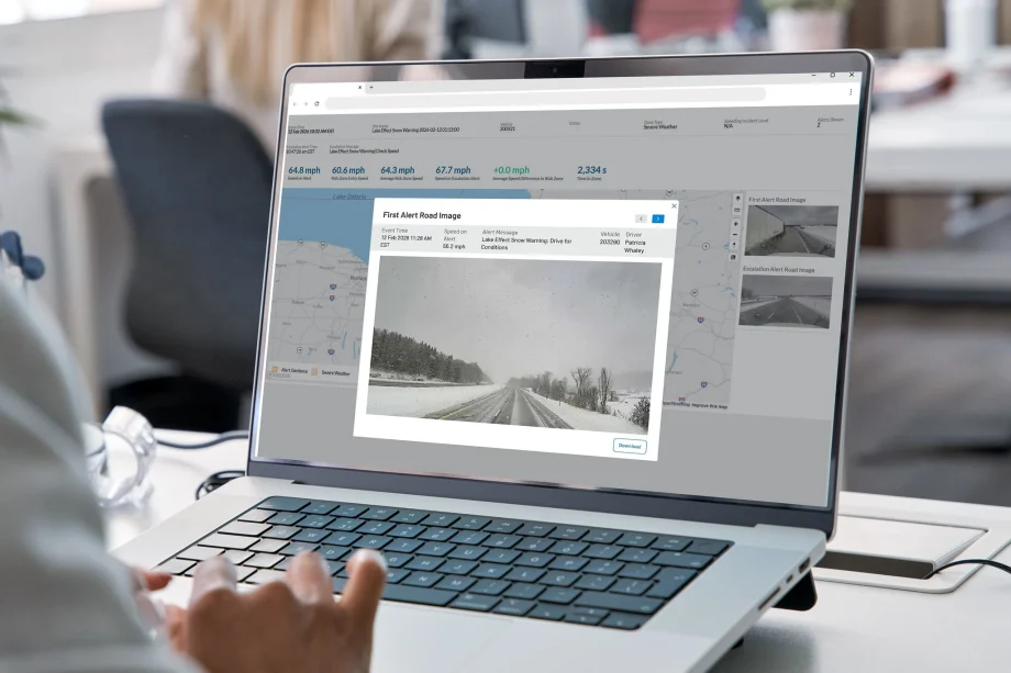 Person using a laptop to view a fleet management dashboard displaying weather alerts, vehicle speed data, maps, and road-condition images. The screen highlights a snowy roadway and a lake-effect snow warning for a fleet vehicle.