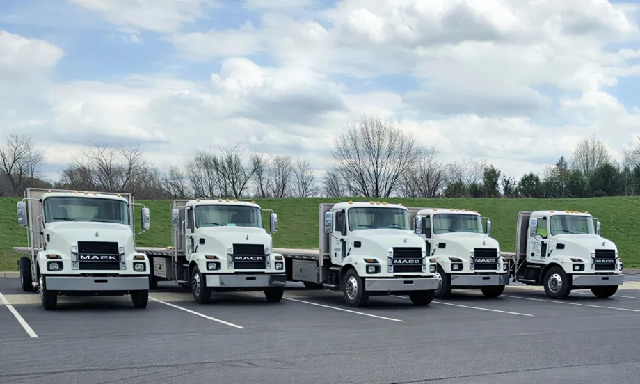 Five white Mack MD-series flatbed trucks are parked side by side in a lot under a cloudy sky, showcasing a fleet of medium-duty commercial vehicles with aluminum flatbed bodies.