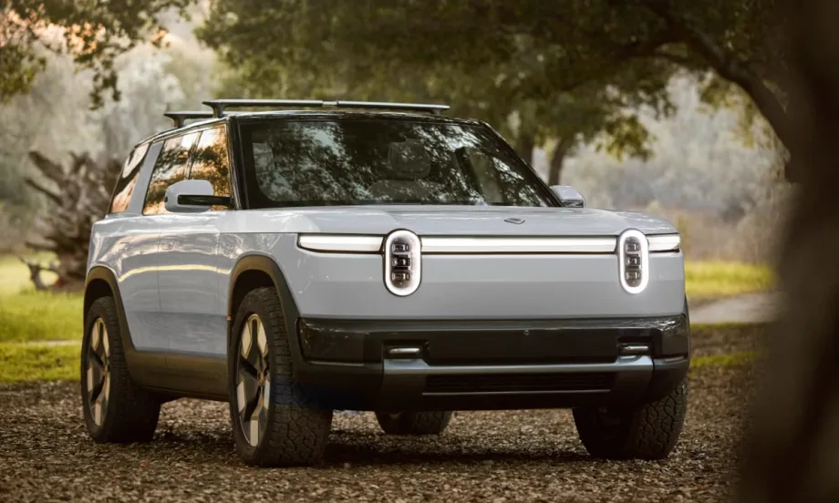 White Rivian R2 electric SUV parked on a leaf-covered path in a wooded area, shown from the front three-quarter angle with distinctive oval headlights, black trim, and roof rails under soft natural light.