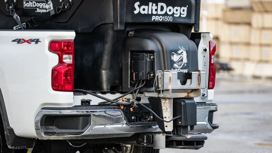 Close-up of a SaltDogg PRO1500 tailgate salt spreader mounted on the rear of a white 4x4 pickup truck, showing the hopper, spreader assembly, wiring connections, and mounting hardware.