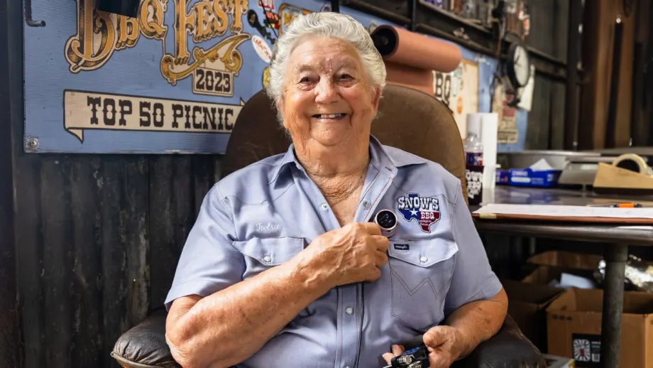 Tootsie Tomanetz, an elderly woman wearing a Snow's BBQ work shirt, smiles while holding a heartbeat recording device inside the barbecue restaurant where she works in Texas.
