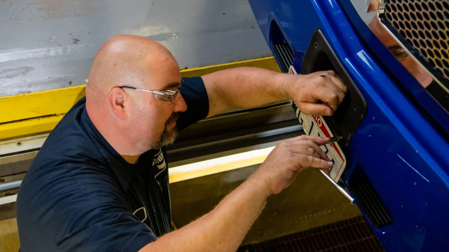 Technician wearing safety glasses inspects a front-mounted ADAS radar sensor on a blue commercial truck.