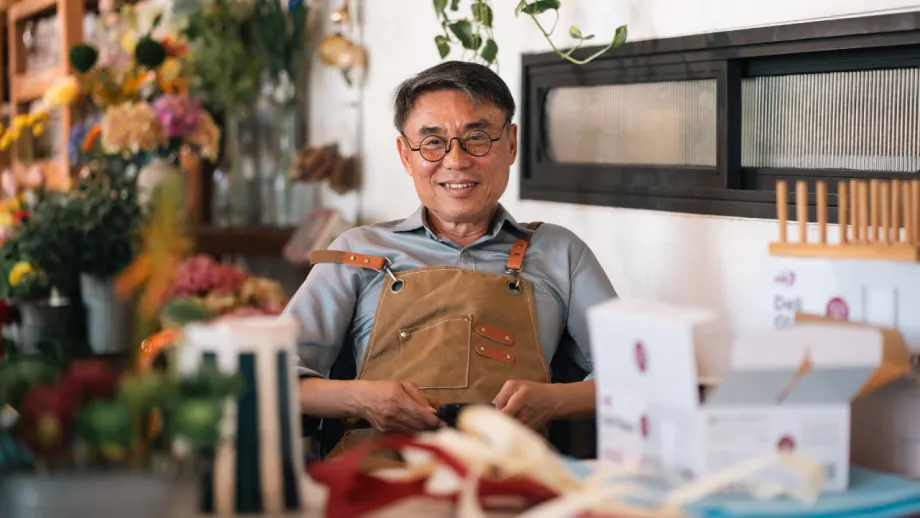 Flower shop owner wearing glasses and an apron sits behind a worktable surrounded by flowers, packaging supplies, and small boxes.