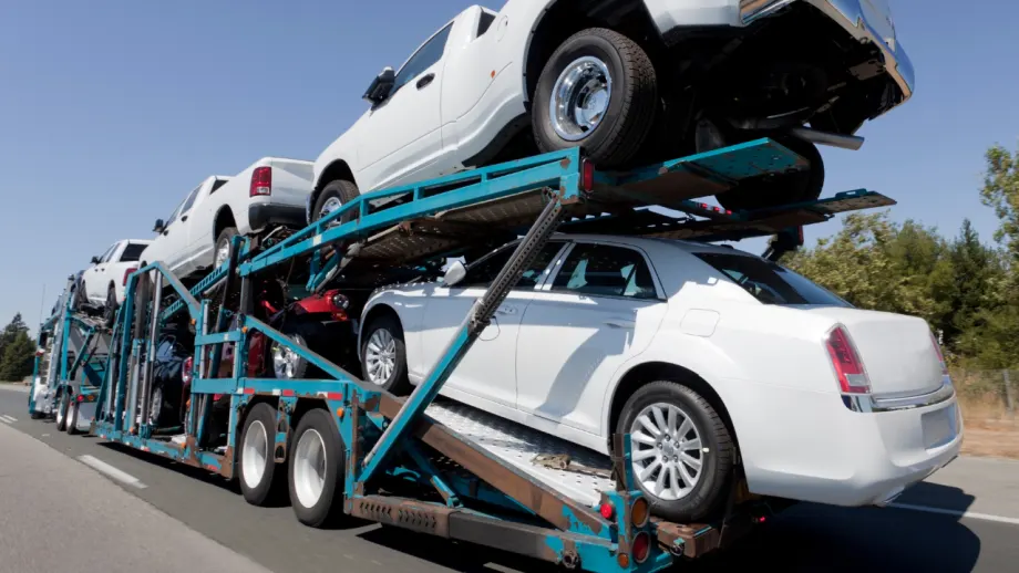 Multi-level car carrier transporting white pickup trucks and sedans along a highway, illustrating commercial vehicle logistics and fleet vehicle delivery.