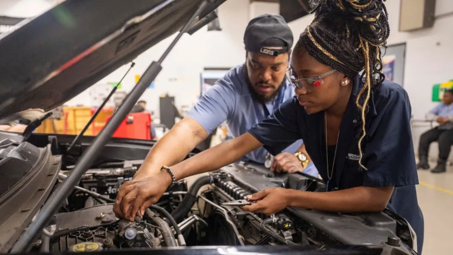 An automotive instructor guides a student wearing safety glasses as they work together under the hood of a vehicle during a hands-on technician training session in a classroom workshop.