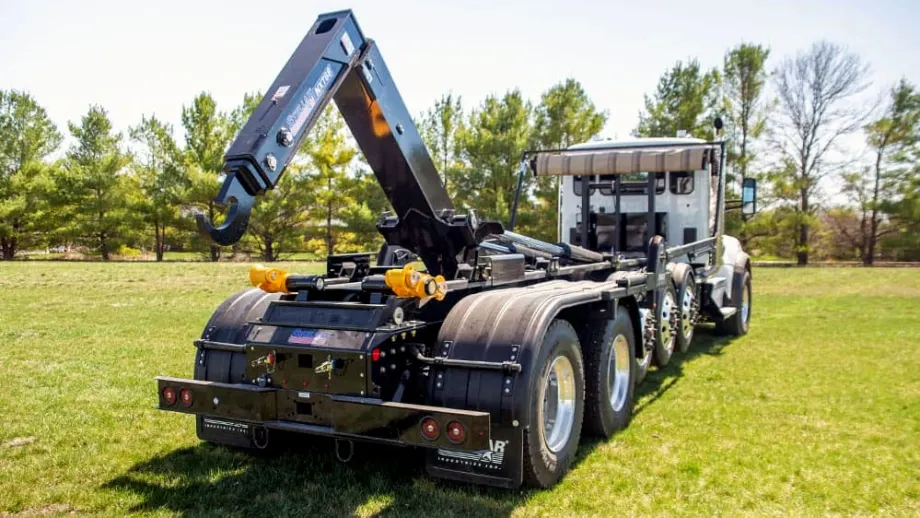 Rear three-quarter view of a heavy-duty truck equipped with a raised hydraulic hooklift system, parked on grass with trees in the background.